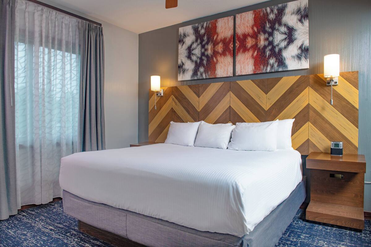 A hotel room with a large bed, two bedside tables, a wall-mounted lamp, and a large window with curtains.
