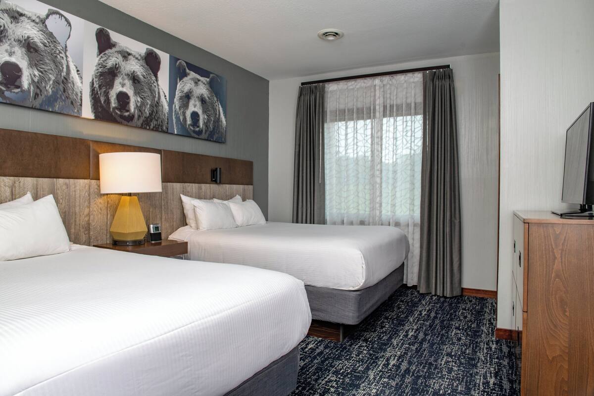 A hotel room with two beds, a wooden headboard, a lamp, a television, and a large window with curtains.