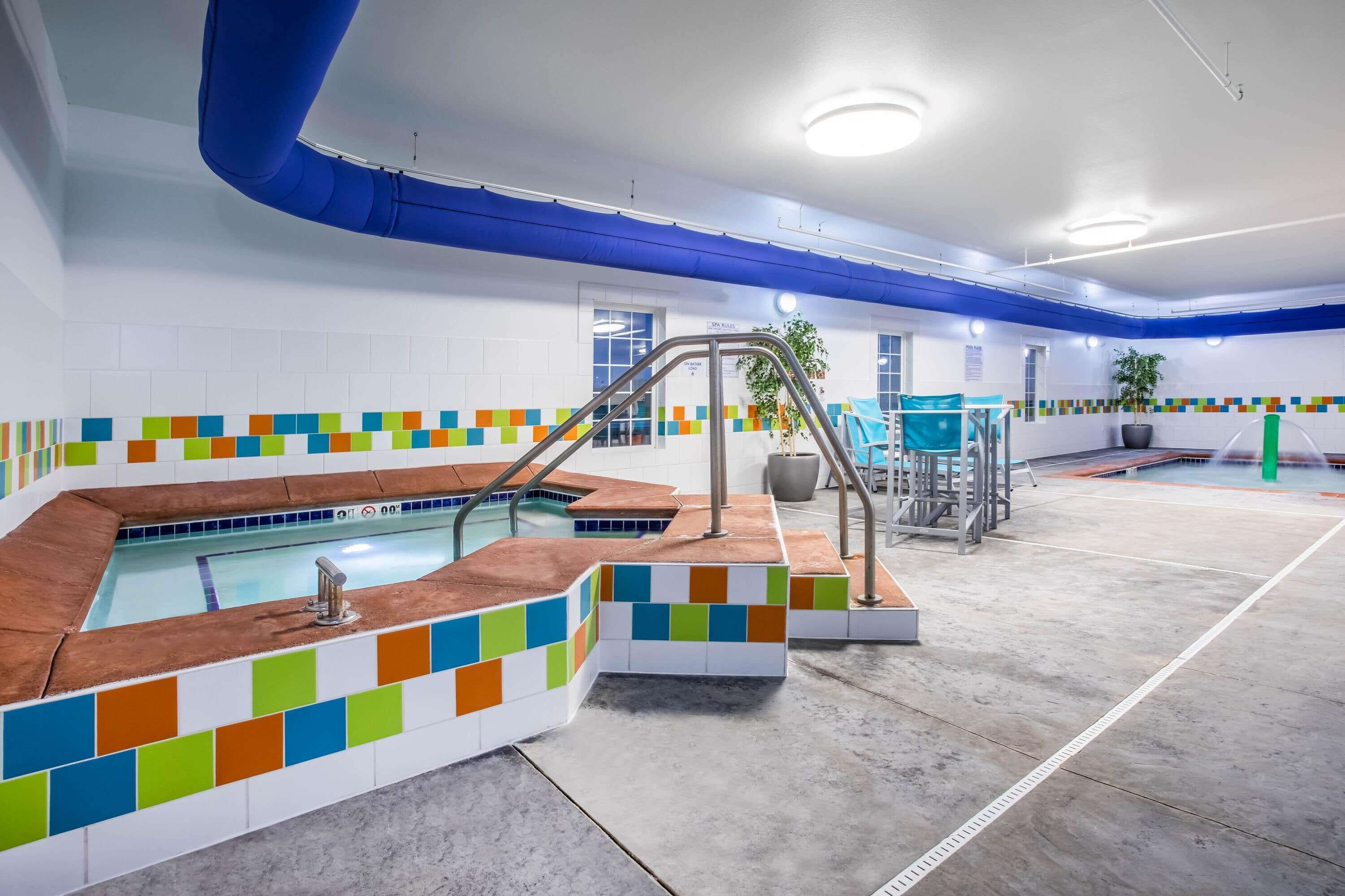 A modern indoor pool area with a colorful tiled border, a central pool, and a seating area with a potted plant.