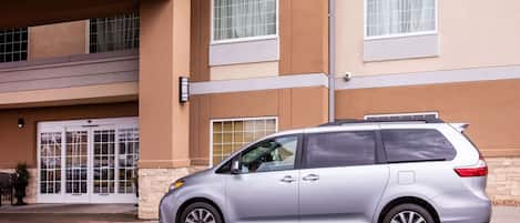 A silver minivan parked in front of a building with a double door entrance, a small balcony, and a window with a curtain.