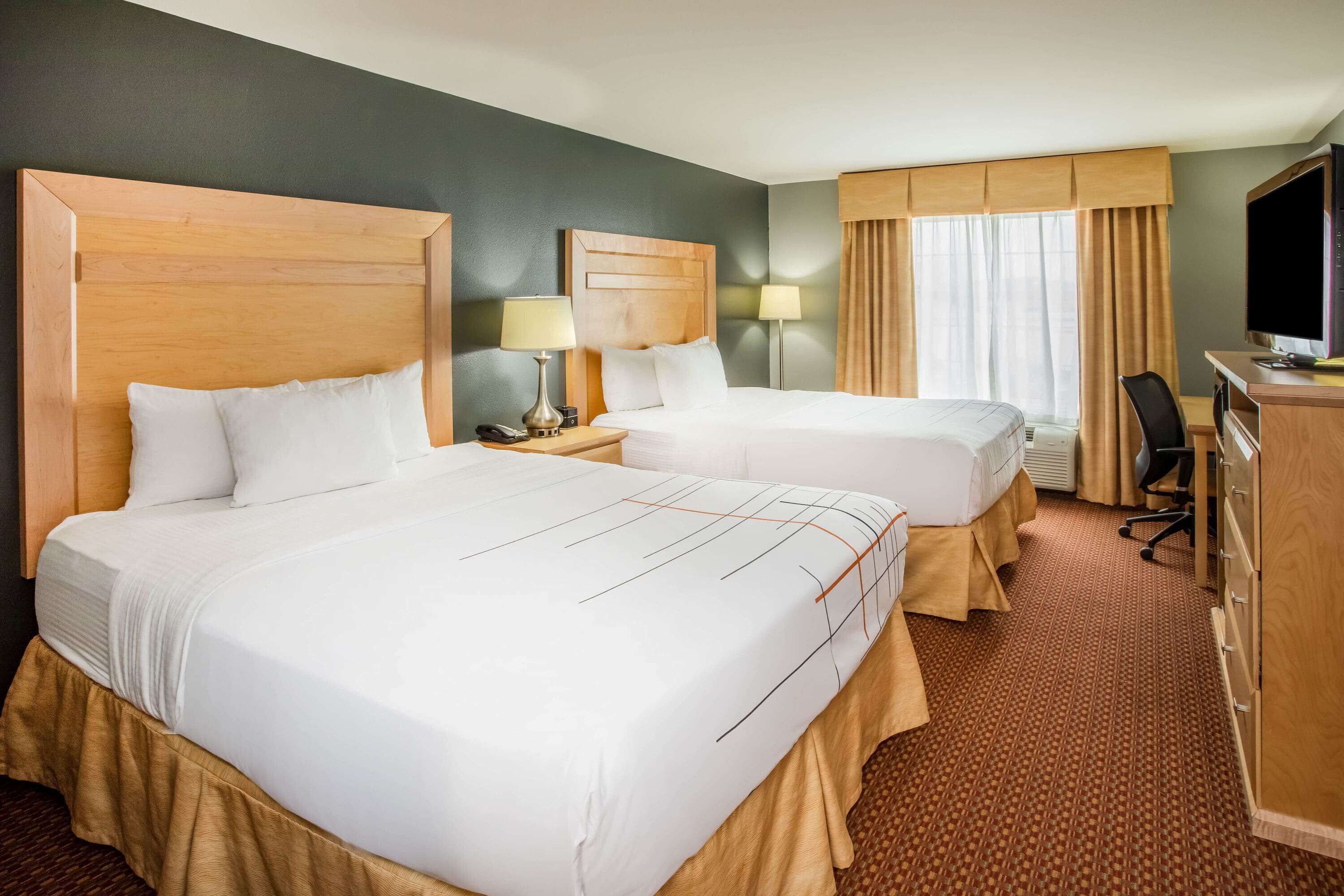 A hotel room with two beds, a desk, a chair, a TV, and a lamp.