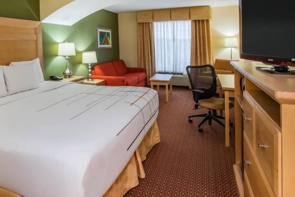 A hotel room with a large bed, a desk, a chair, a red sofa, a television, and a window with curtains.