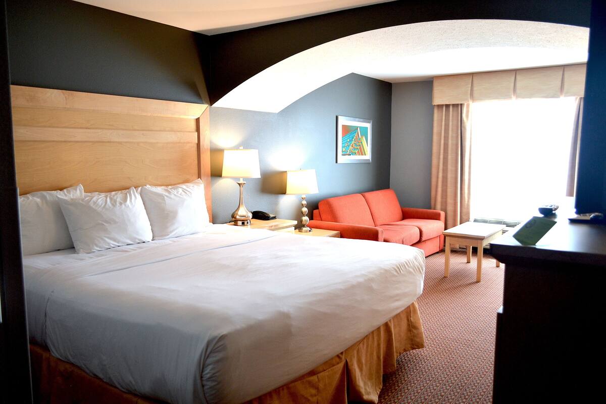 A hotel room with a large bed, a desk, a chair, a sofa, and a window with curtains.
