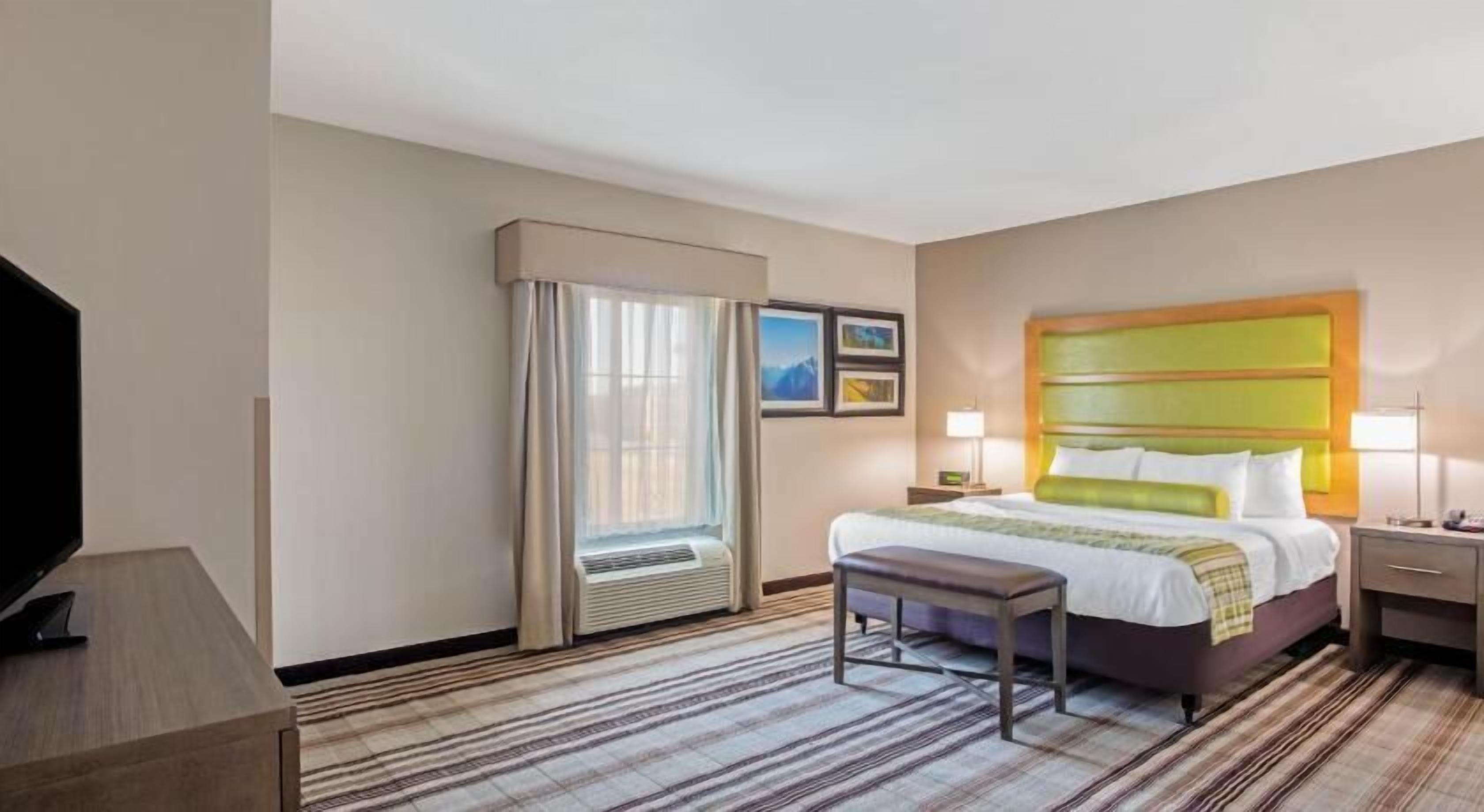 Ein Hotelzimmer mit Bett, Schreibtisch, Fernseher, einem Fenster mit Vorhängen und einer Wand-Klimaanlage.