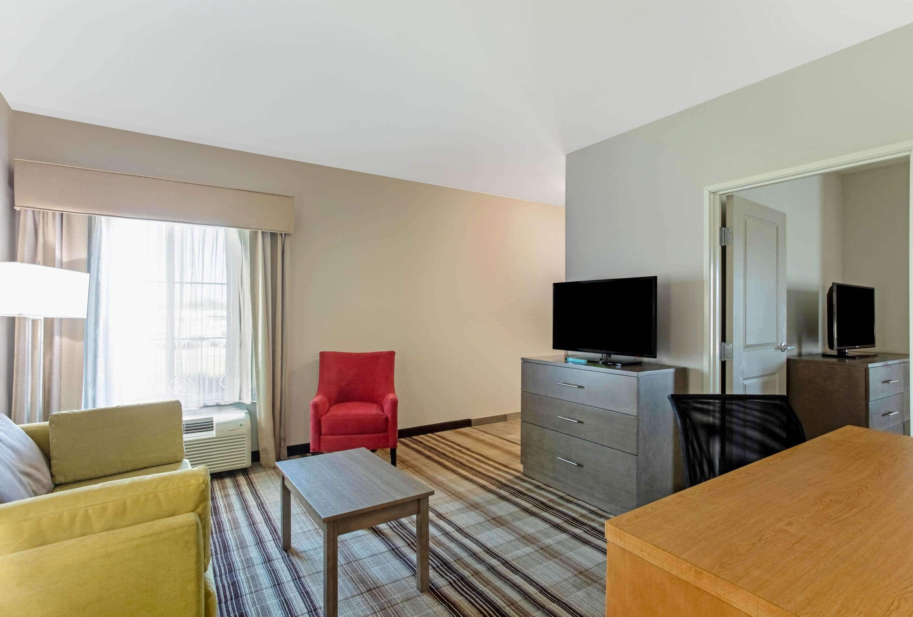A hotel room with a sofa, a red armchair, a wooden table, a TV on a dresser, and a mirror on the wall.