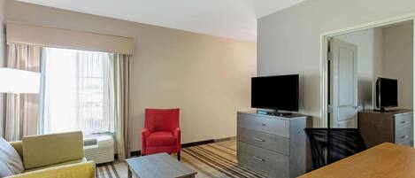 A hotel room with a sofa, a red armchair, a wooden table, a TV on a dresser, and a mirror on the wall.