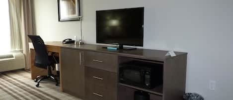A hotel room with a desk, chair, television, microwave, and a trash bin.
