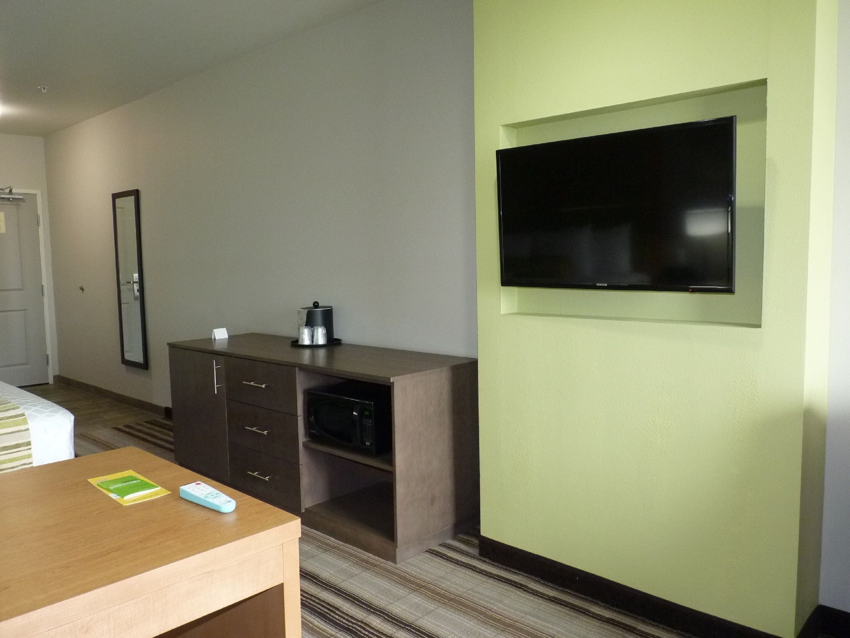 A hotel room with a bed, a desk with a remote control, a coffee maker, a TV mounted on the wall, and a door.