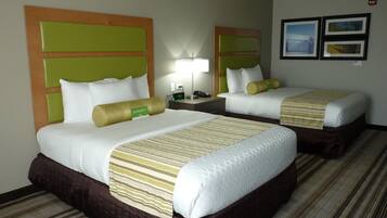 A hotel room with two beds, each with a green headboard, white bedding, and a striped blanket. There is a nightstand with a lamp and a clock between the beds.