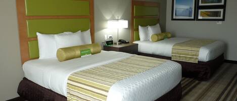 A hotel room with two beds, each with a green headboard, white bedding, and a striped blanket. There is a nightstand with a lamp and a clock between the beds.