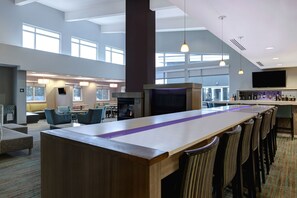 A spacious hotel lobby with a long reception desk, multiple seating areas, a fireplace, and a bar with a television.