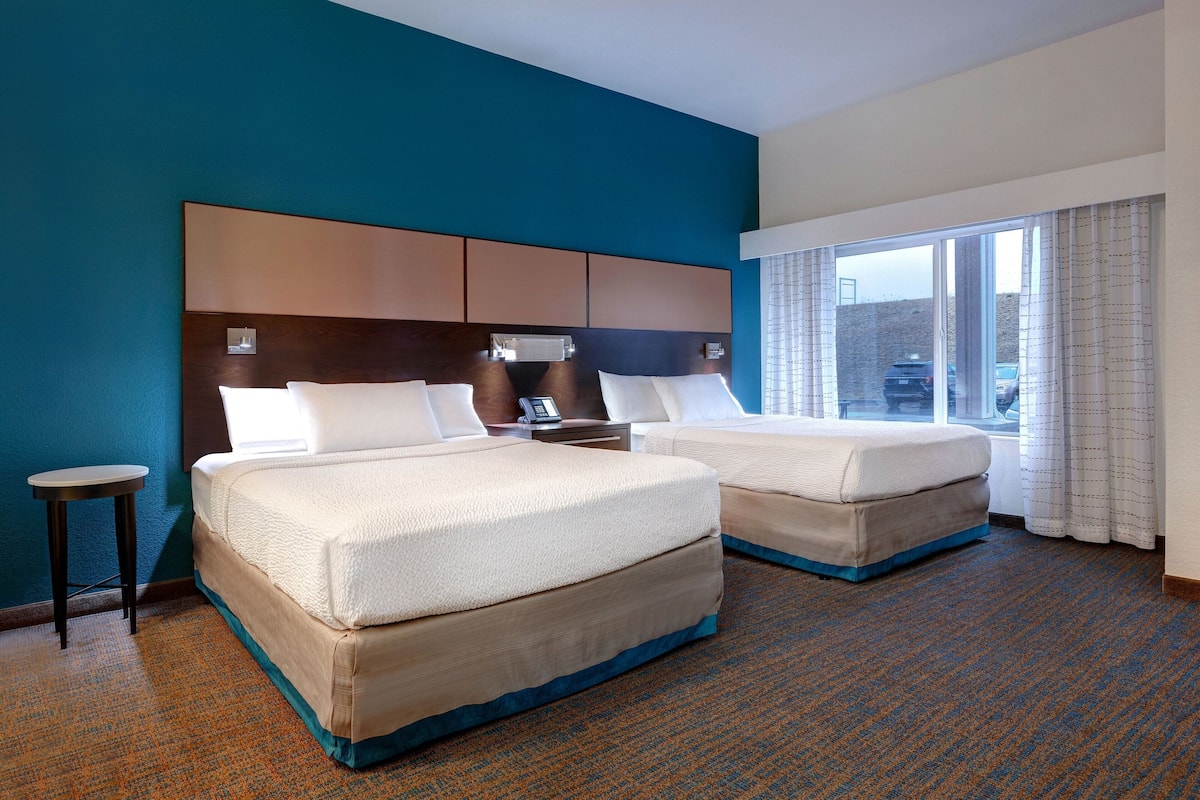 A hotel room with two beds, a small table, and a large window with curtains.