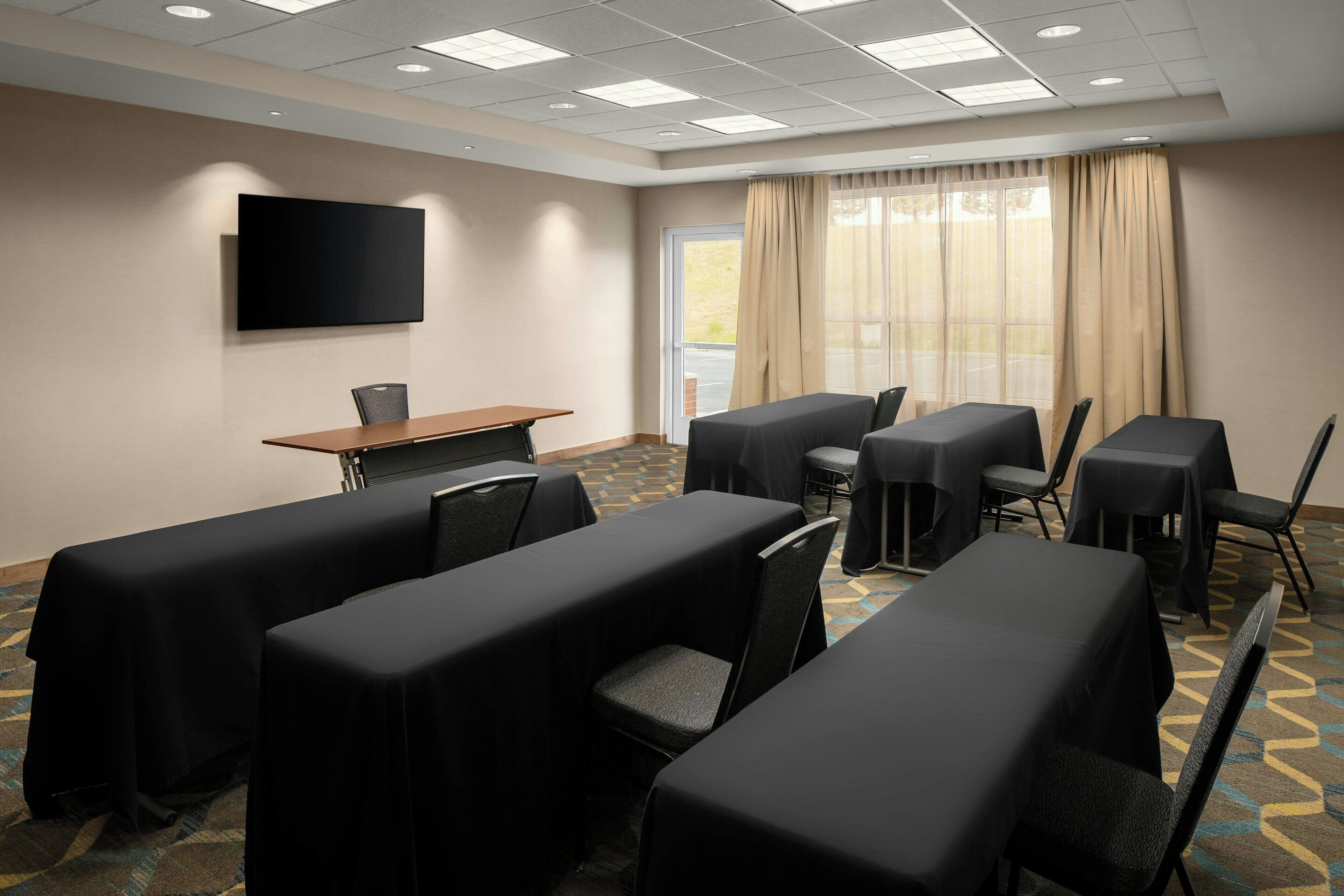 A conference room with tables covered in black tablecloths, a flat-screen TV mounted on the wall, and a large window with beige curtains.