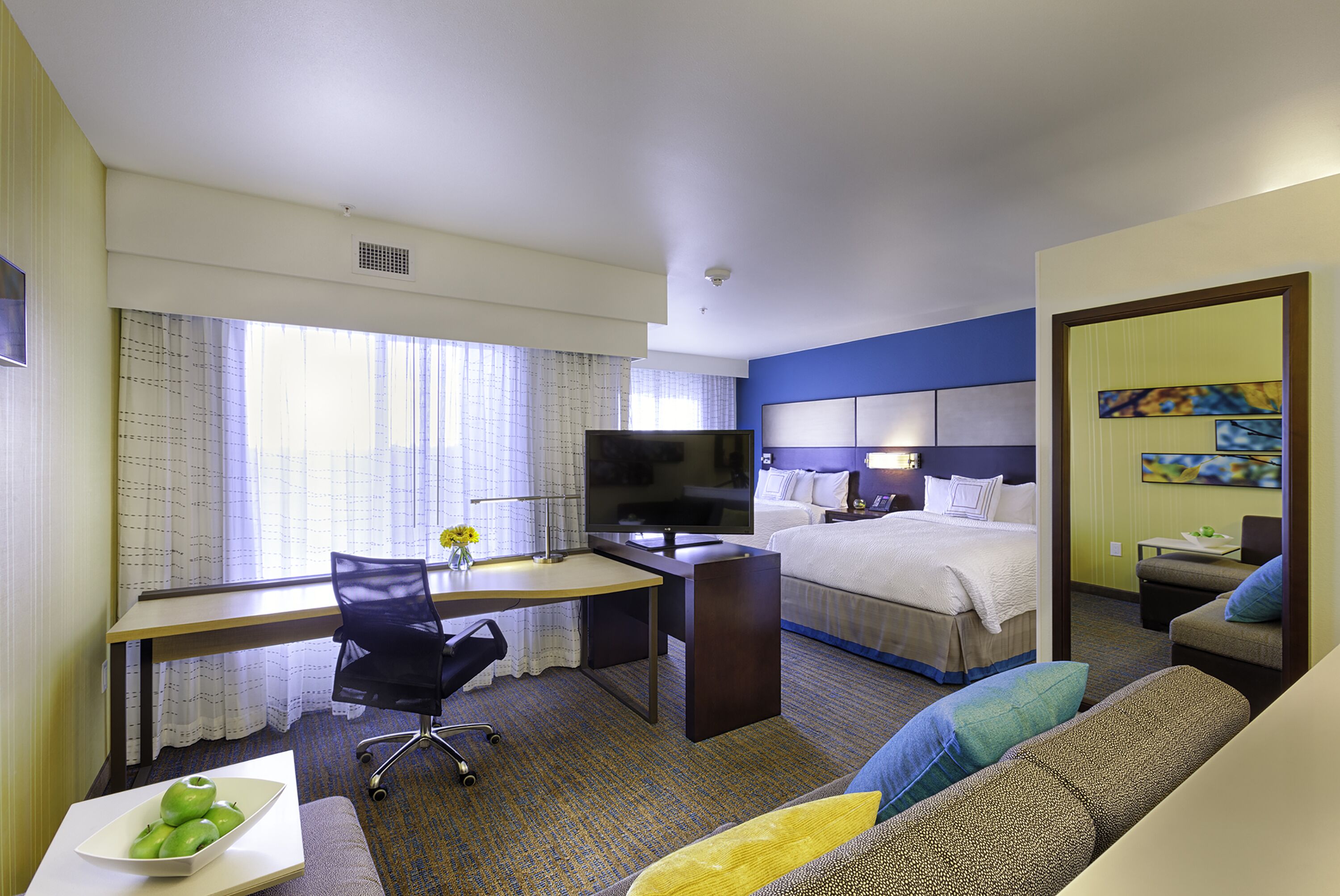 Une chambre d’hôtel comprenant un lit, un bureau avec un ordinateur, une télévision, un canapé et une table à manger avec un saladier de fruits.