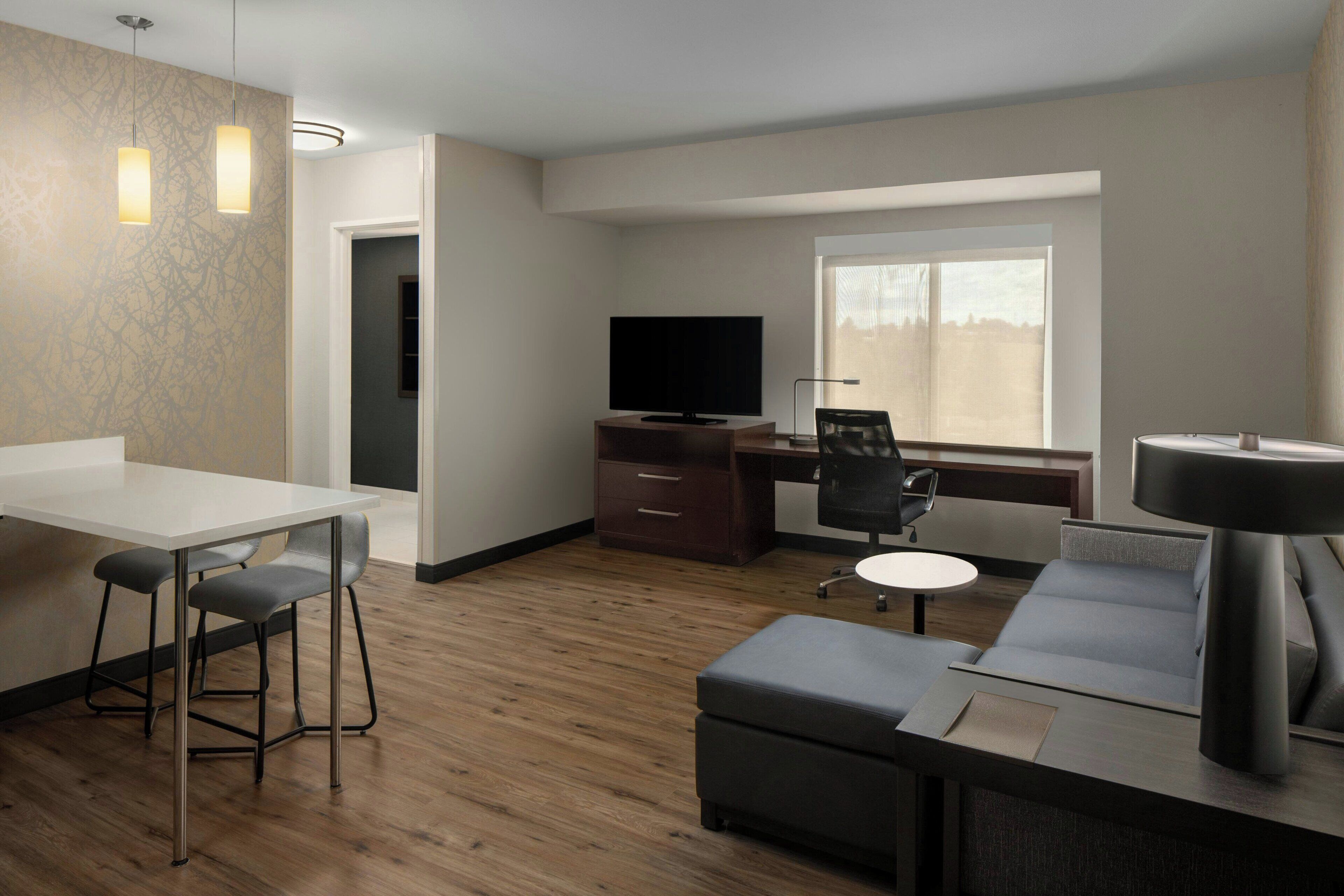 Une chambre d’hôtel moderne avec un plancher en bois, un coin repas comprenant une table et des chaises, un canapé, un bureau avec un ordinateur, une télévision et une grande fenêtre laissant entrer la lumière naturelle.