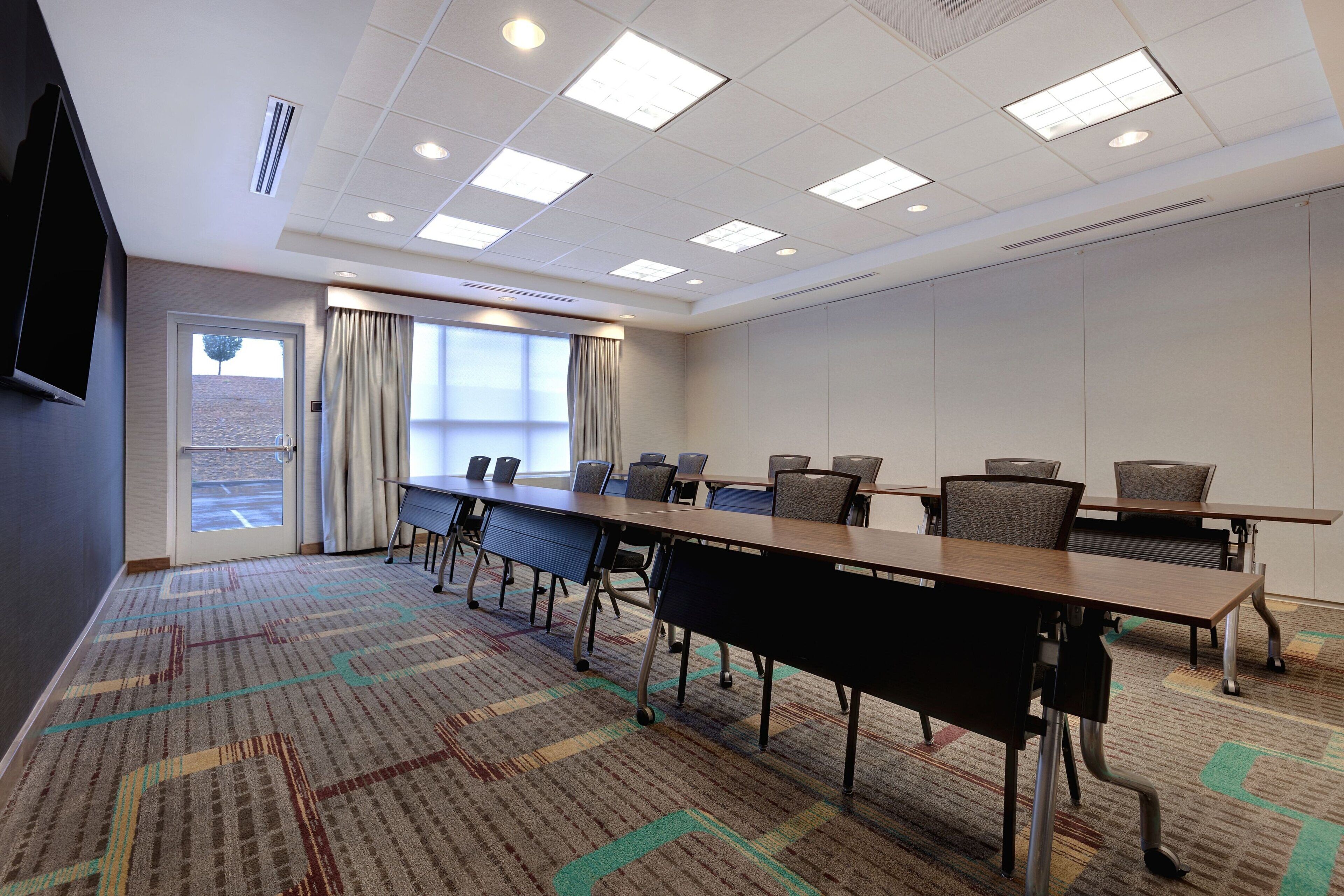 A conference room with a long table, chairs, a television, and a window with curtains.