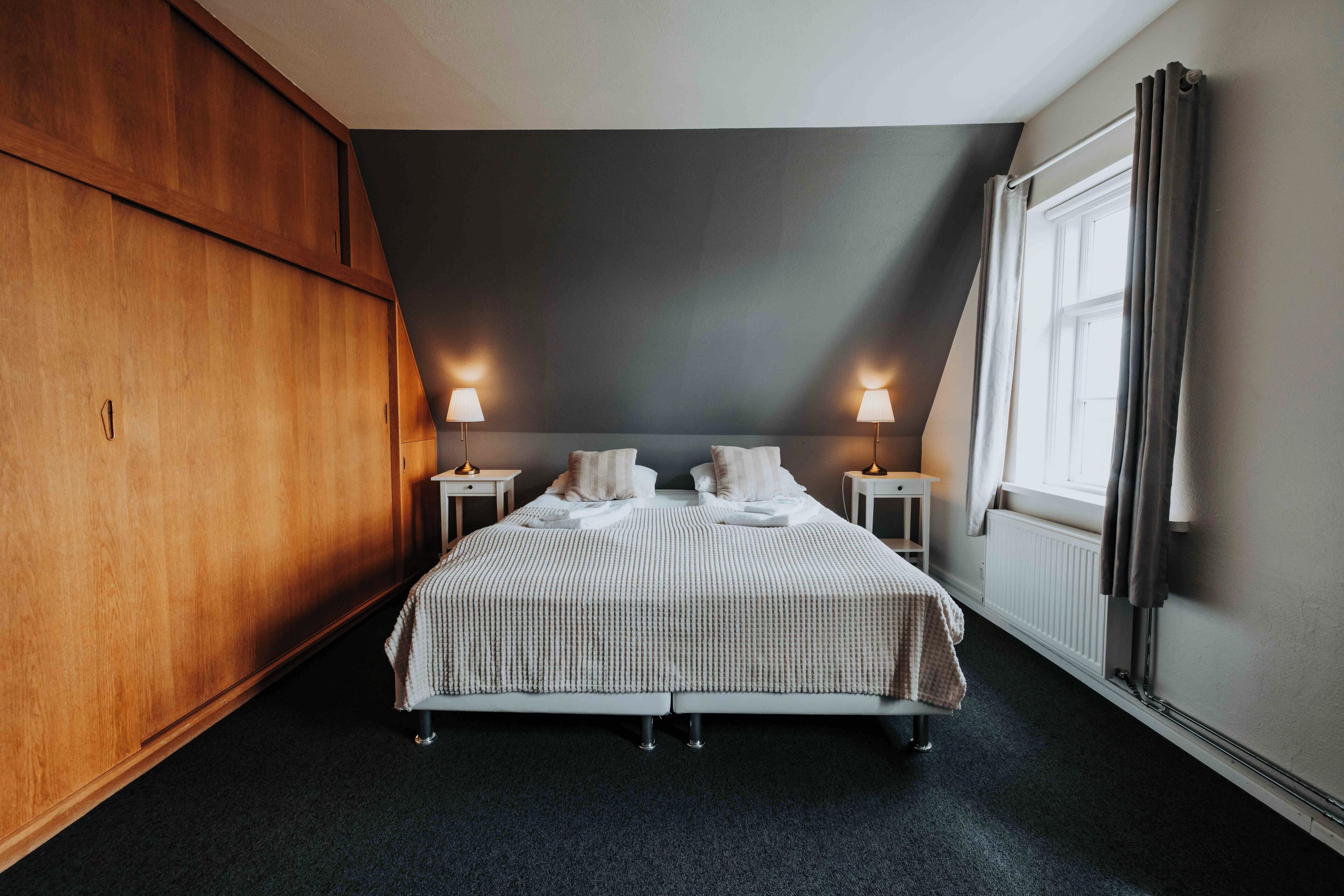 Una habitación de hotel con una cama grande, dos lámparas de noche, un ventanal con cortinas y un armario de madera.