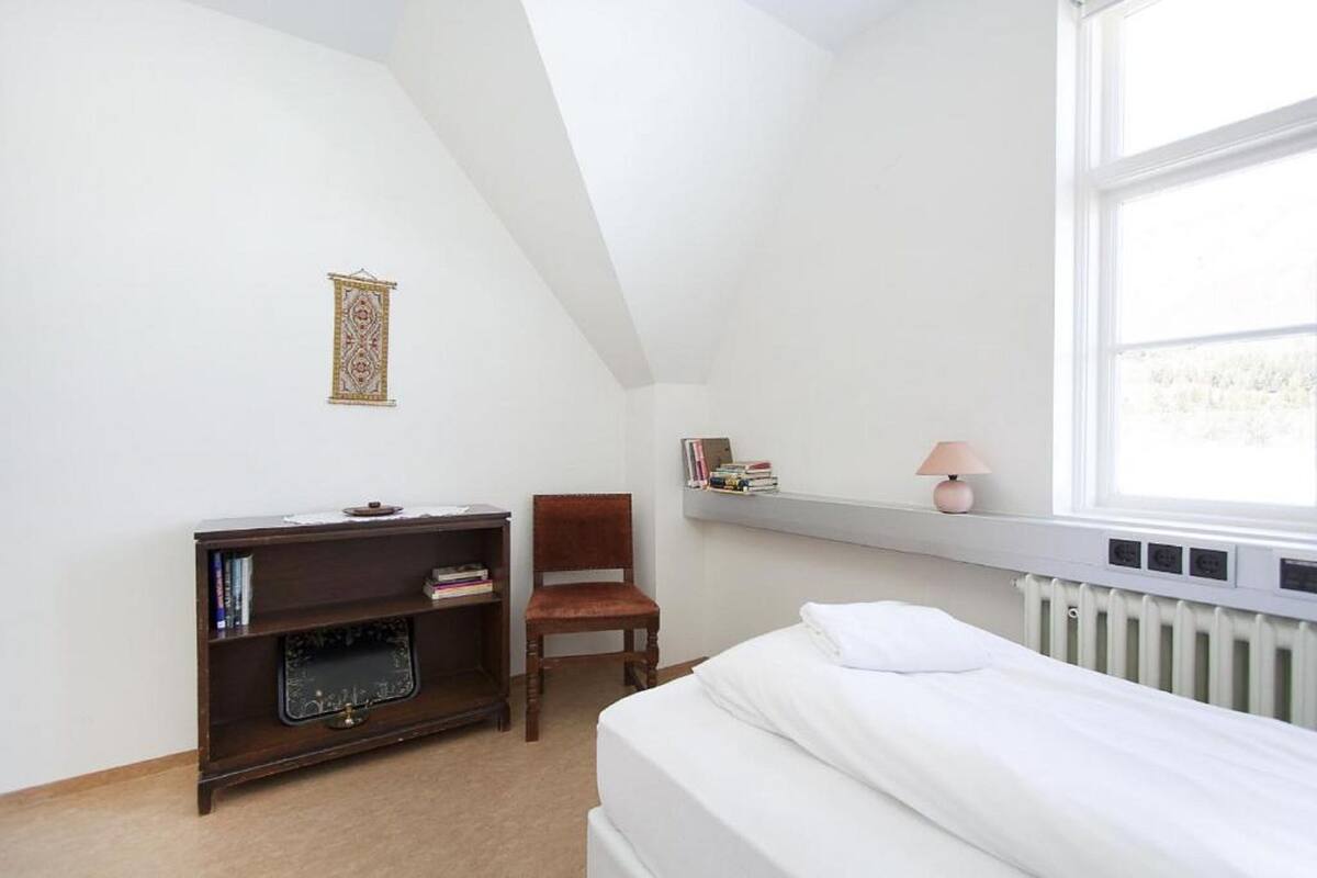 A bedroom with a bed, a bookshelf, a chair, a radiator, and a window.