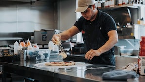 Una persona preparando comida en una cocina con electrodomésticos de acero inoxidable y estantes llenos de condimentos.