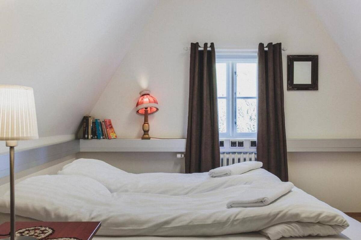 A bedroom with a bed, a bedside lamp, a window with curtains, a radiator, and a shelf with books.