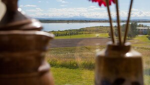 Una ventana con vistas a un lago y montañas lejanas, un florero con flores, y un frasco decorativo.
