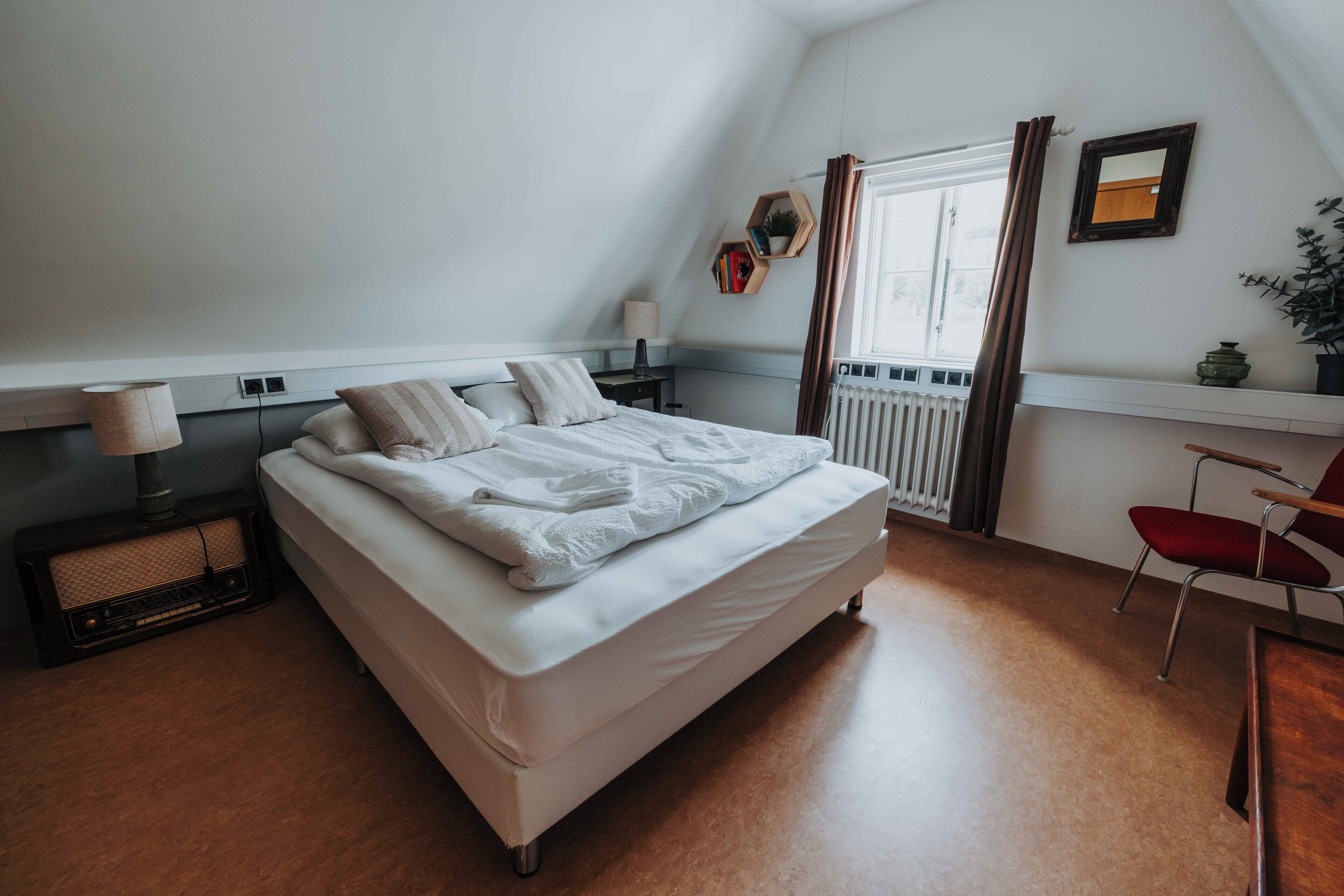 Un dormitorio con cama, mesita de noche, lámpara, ventana con cortinas, radiador, silla y estante con una planta.