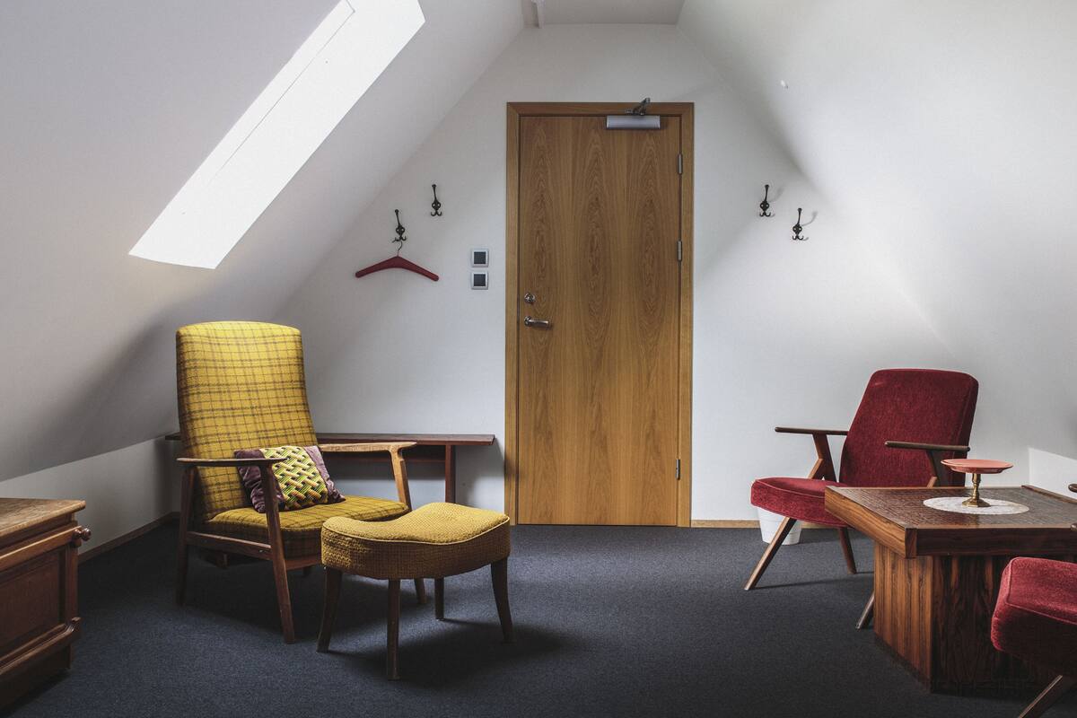 A room with a sloped ceiling, a wooden door, a red chair, a yellow chair, a small table, and a hanging coat rack.