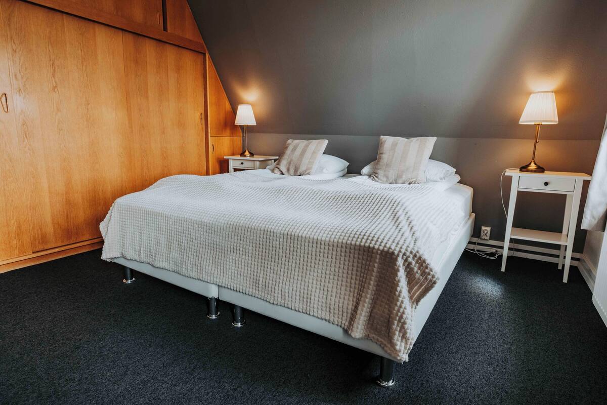 A neatly made bed with a white comforter and two pillows, flanked by two bedside tables with lamps, in a room with dark carpeting and wooden paneling.
