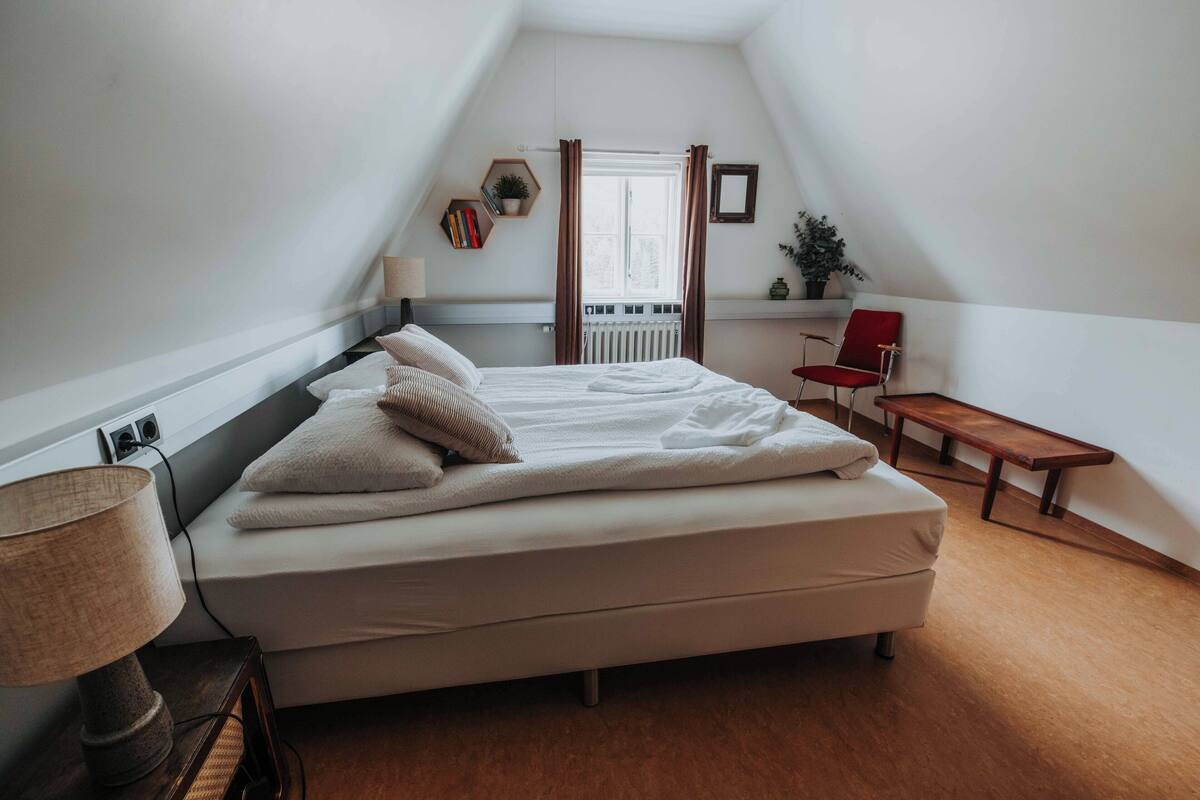 A bedroom with a bed, a bedside table with a lamp, a red chair, a wooden bench, and a window with curtains.