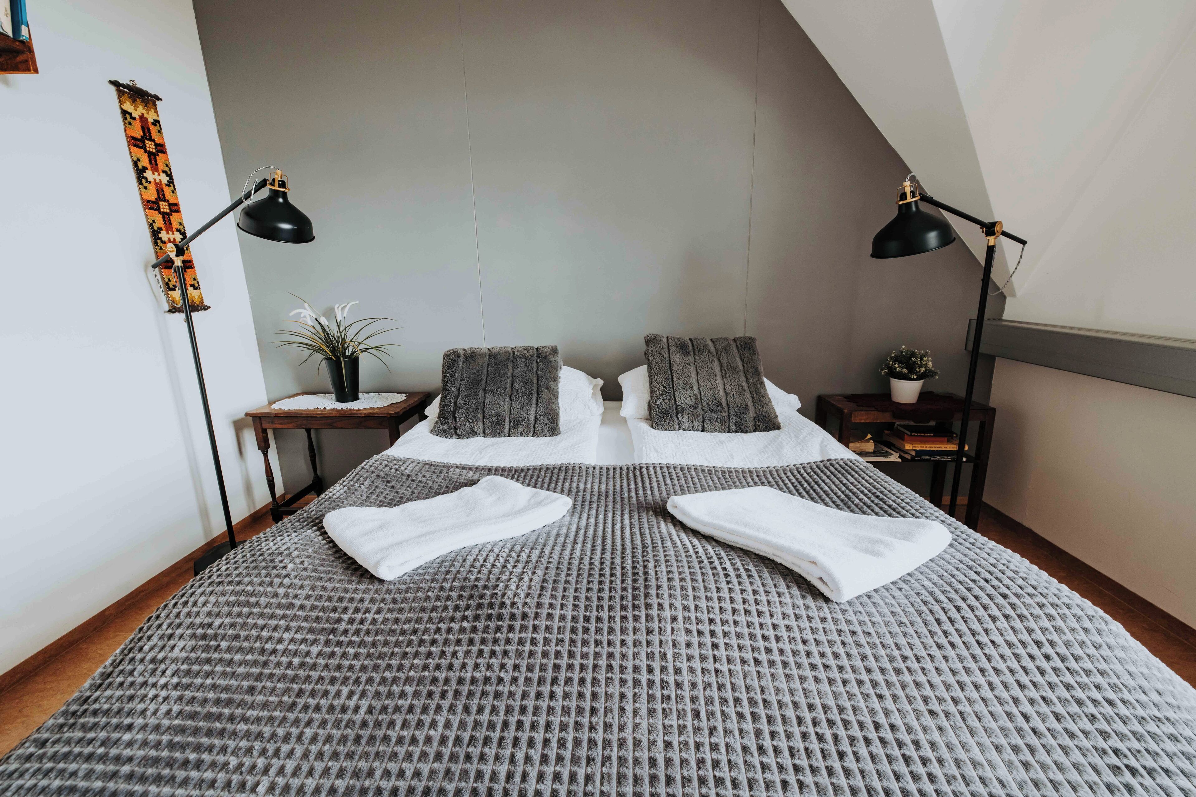 A neatly made bed with a grey textured bedspread, two pillows, and two white towels folded on top. There are two black floor lamps on either side of the bed, a small wooden nightstand with a potted plant, and a wall-mounted shelf with books.