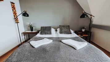 A neatly made bed with a grey textured bedspread, two pillows, and two white towels folded on top. There are two black floor lamps on either side of the bed, a small wooden nightstand with a potted plant, and a wall-mounted shelf with books.