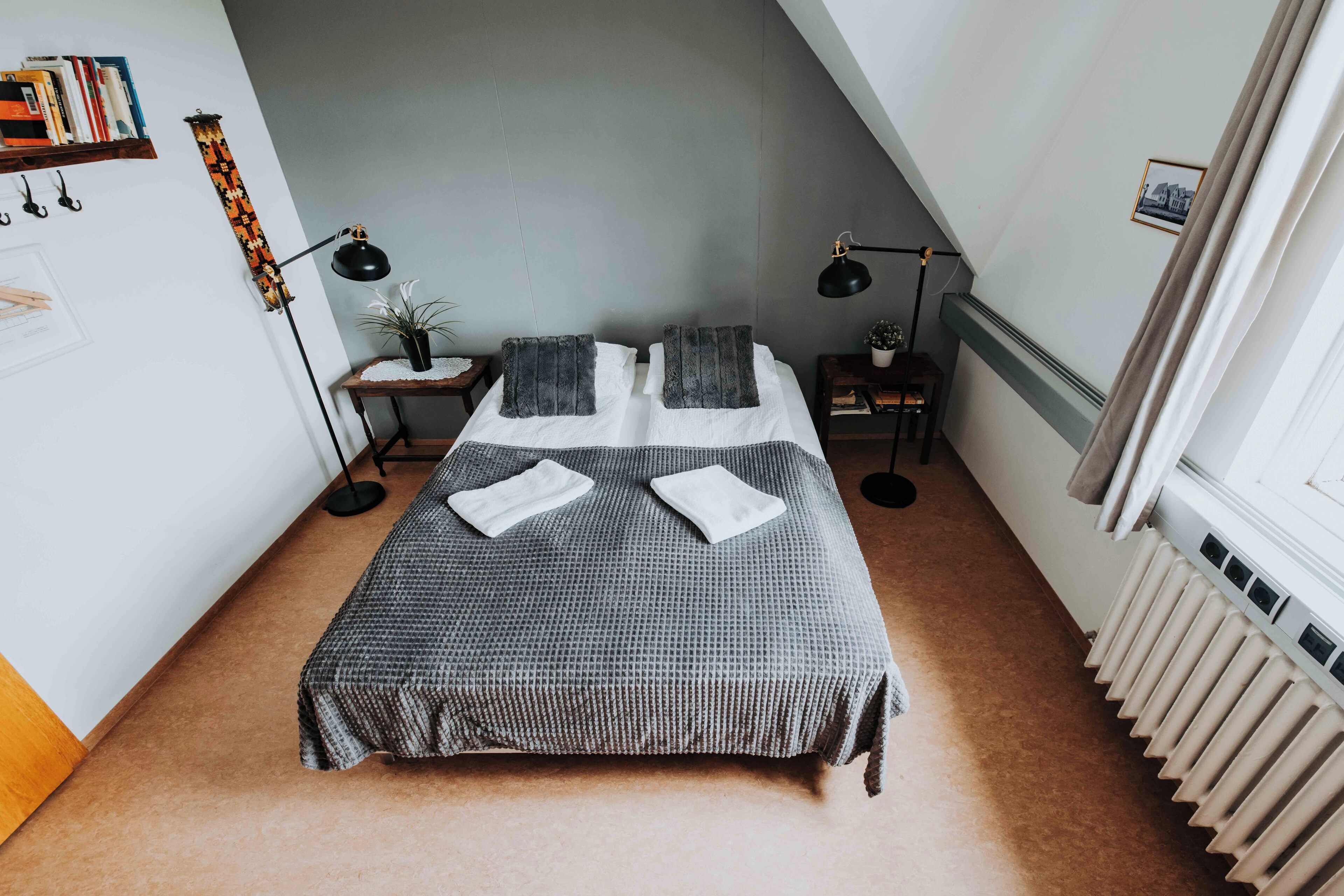 A neatly arranged bedroom with a bed, two bedside lamps, a small table with a plant, and a radiator.