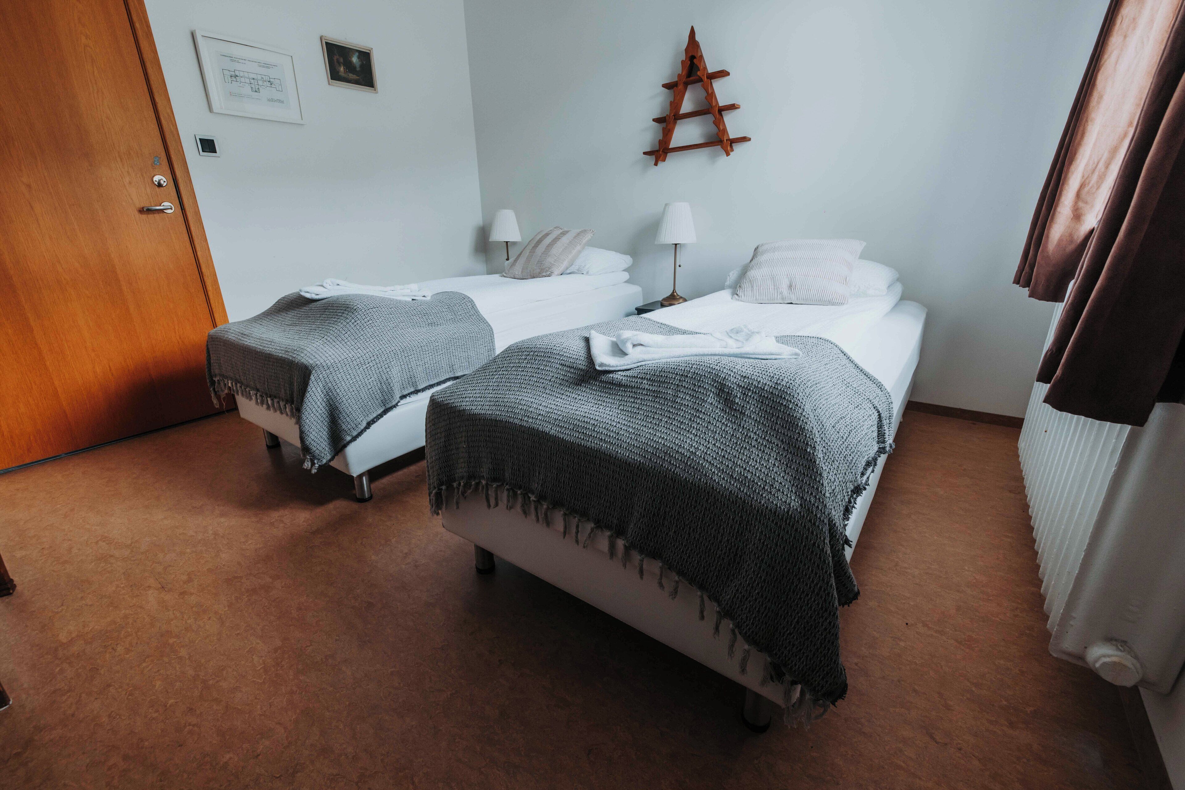 A room with two single beds, each with a bedside table and lamp, a wooden ladder on the wall, and a radiator on the right.