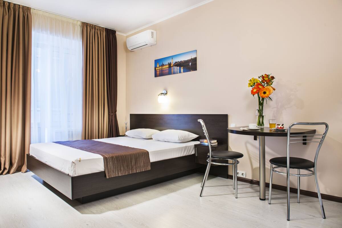 A hotel room with a bed, a desk with two chairs, a vase with flowers, and a picture on the wall.
