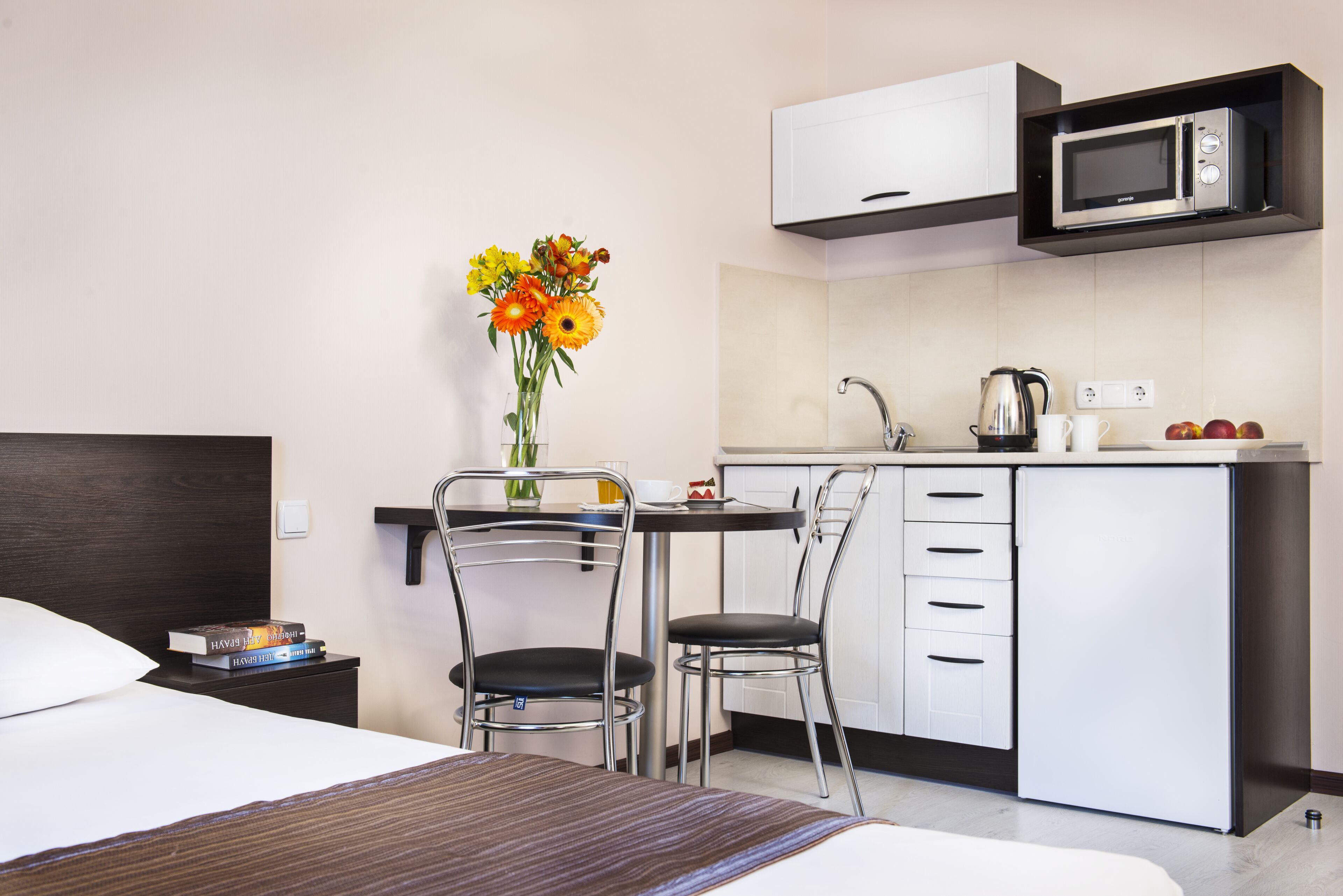 Une chambre d’hôtel moderne équipée d’un lit, d’un bureau avec une chaise, d’un micro-ondes, d’une bouilloire et d’un vase avec des fleurs.