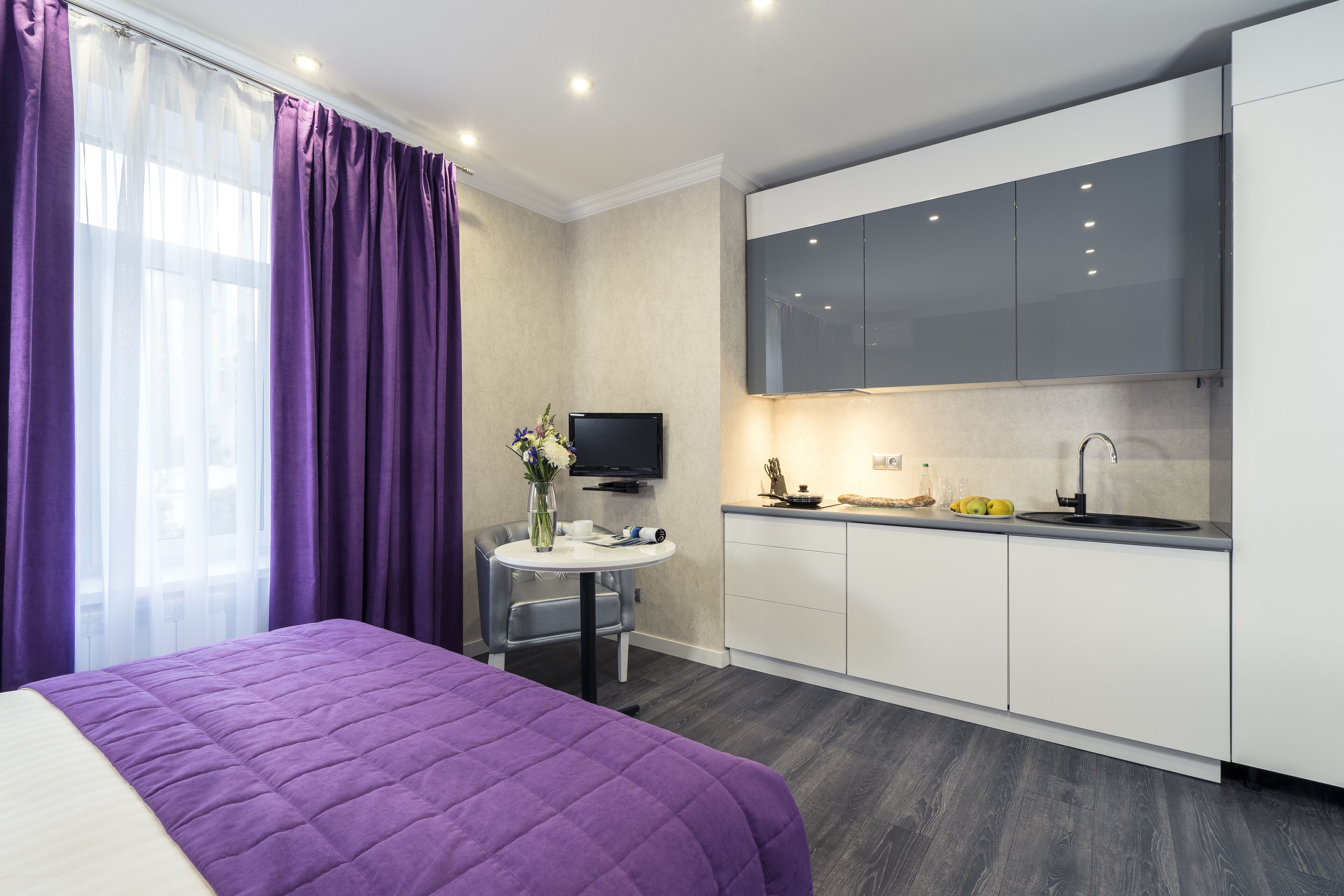 Una habitación de hotel moderna con una cama, una mesita redonda con un florero, un televisor de pantalla plana, una zona de cocina con armarios y un lavabo, y cortinas moradas.