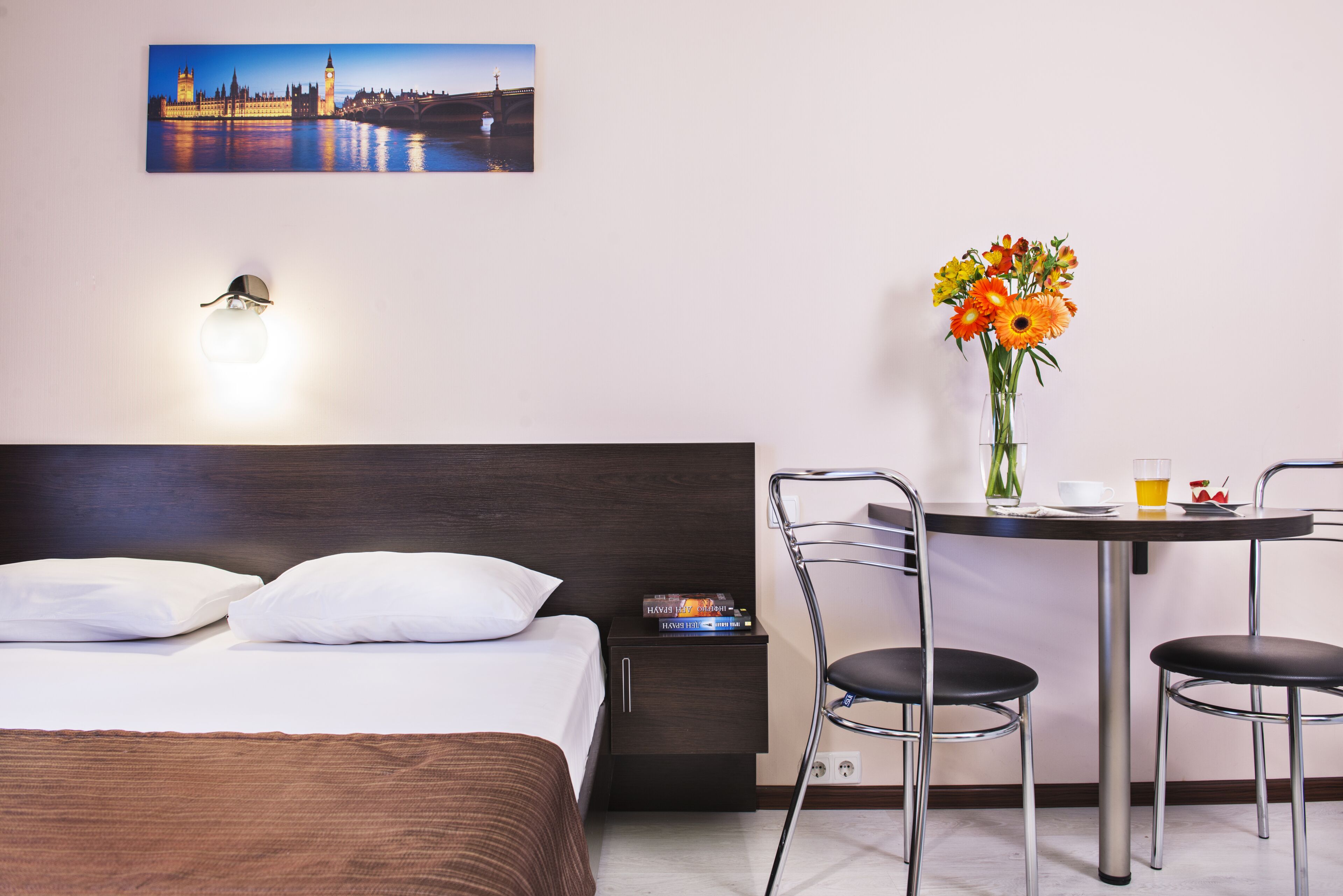 A hotel room with a bed, a nightstand with a lamp, a table with chairs, a vase of flowers, and a painting of a cityscape.