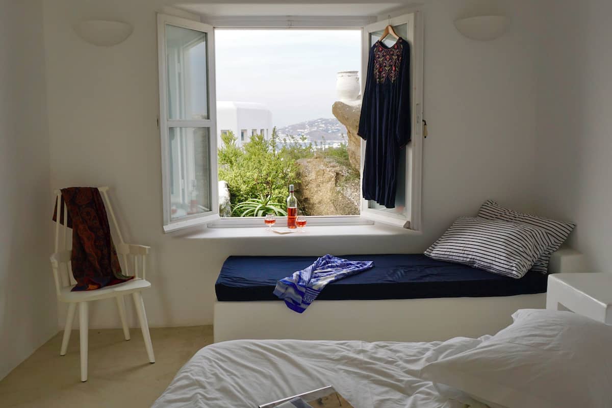 A bedroom with a bed, a window seat with a view, a chair, and a hanging garment.