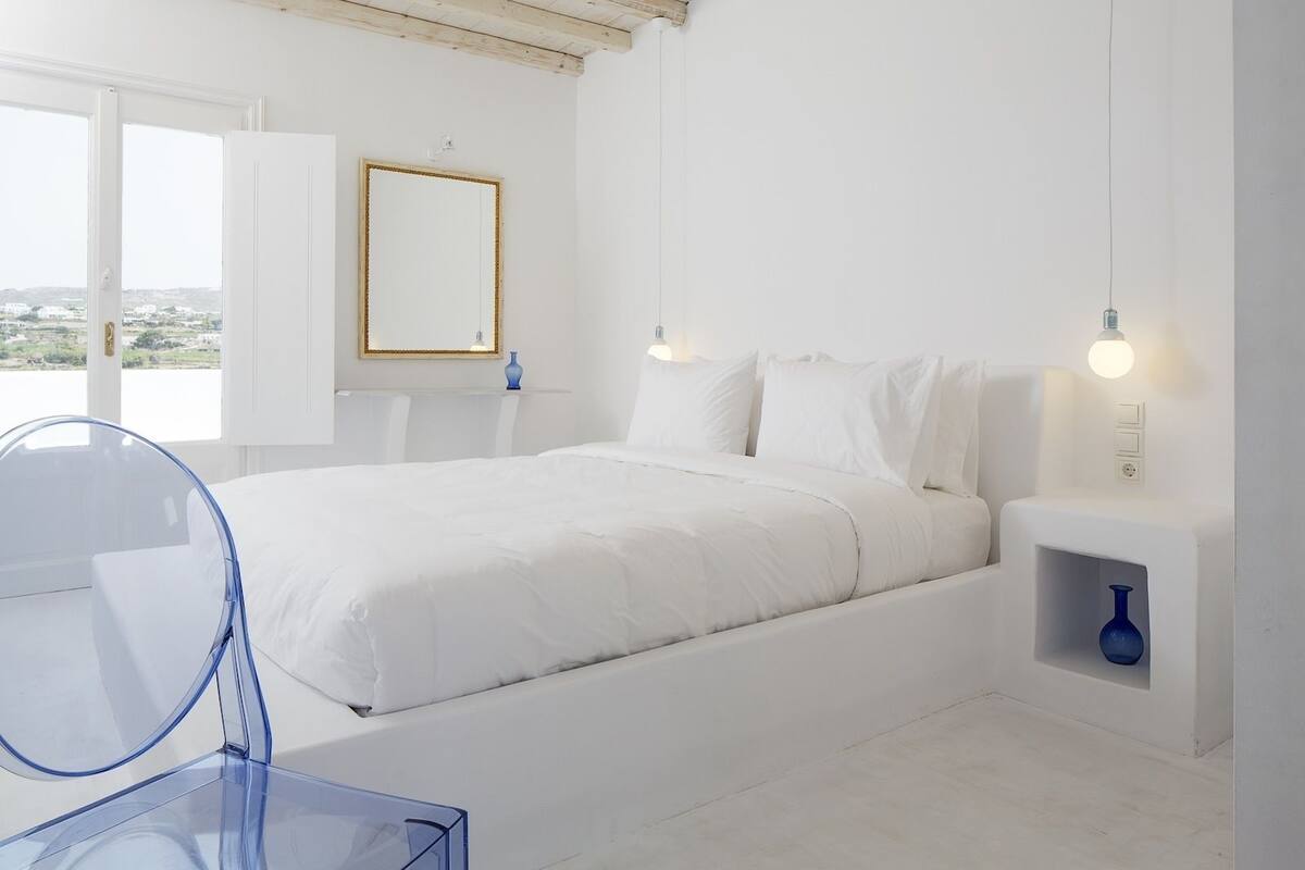 A modern, minimalist bedroom with a large bed, bedside tables, a mirror, and a view of the outdoors.