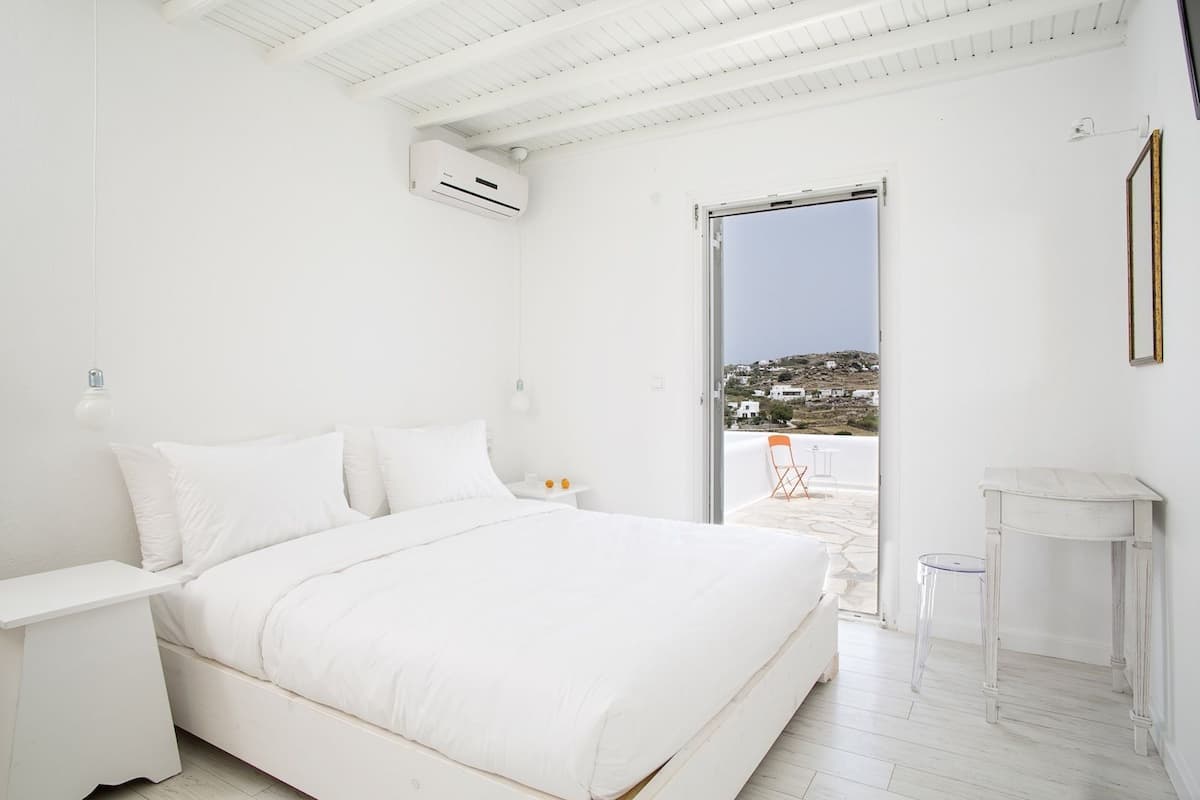 A white bedroom with a bed, bedside table, and an air conditioning unit. There is a balcony with a view of a hillside.