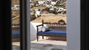 Una cama de madera con colchón azul y almohada a rayas, ubicada en un balcón con suelo de piedra, con vistas a una colina de edificios blancos.