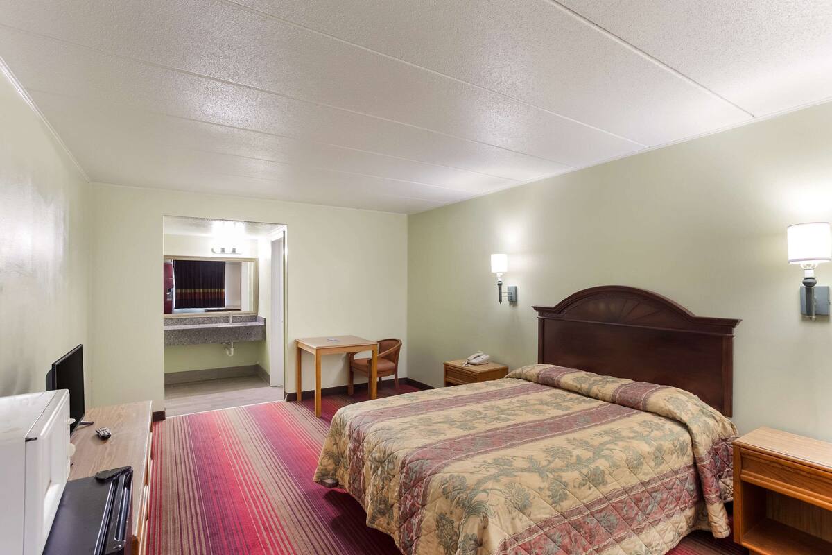 A hotel room with a bed, a desk, a chair, a TV, and a bathroom visible through an open door.