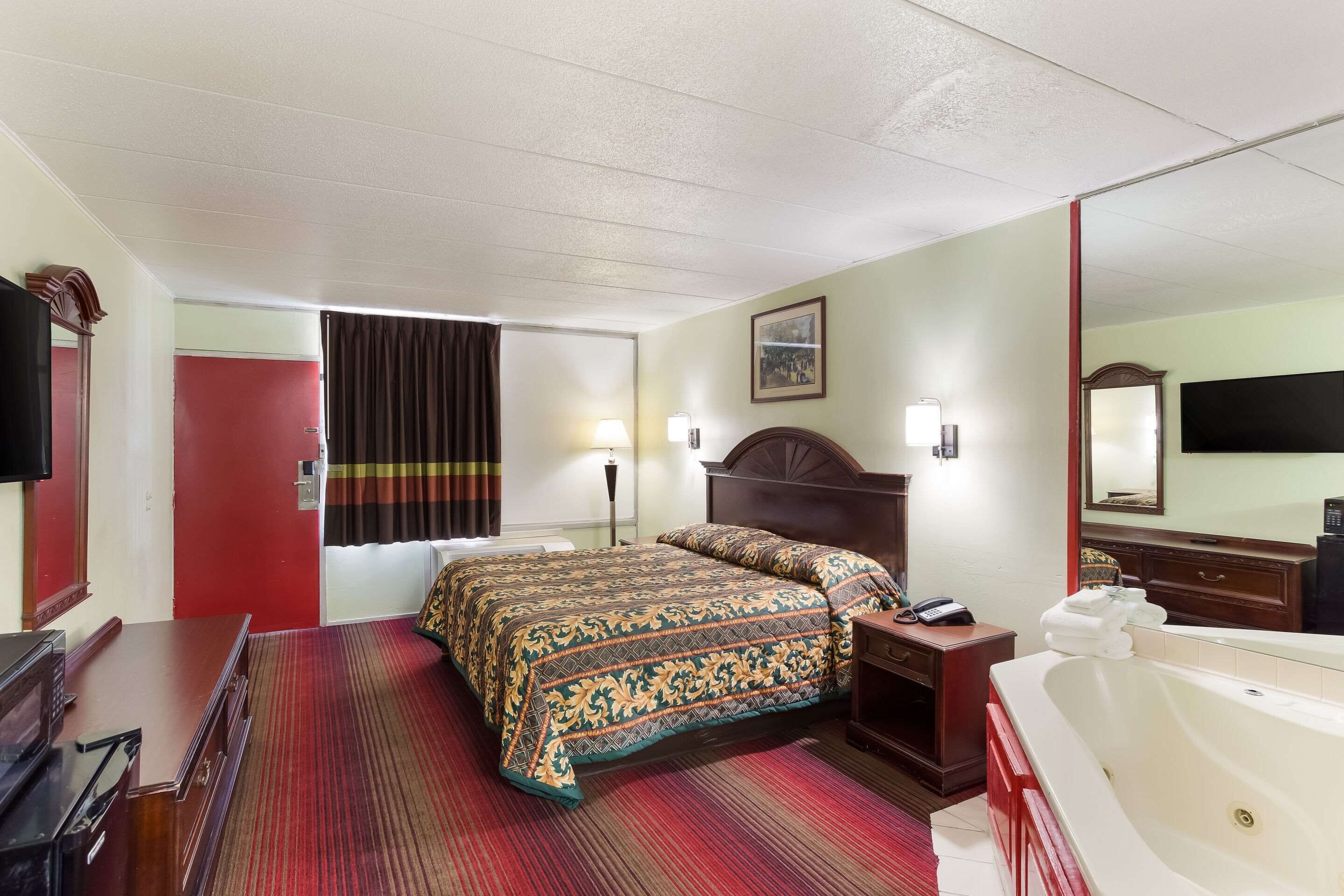 Quarto de hotel com uma cama grande, uma escrivaninha, uma cadeira, uma luminária de mesa, uma mesa de cabeceira, uma TV, um espelho, um banheiro com banheira e uma porta vermelha.