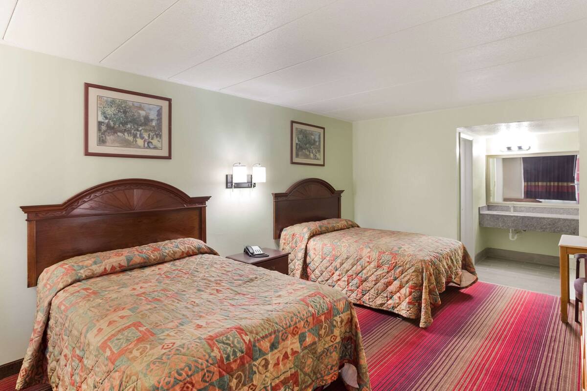 A hotel room with two beds, a nightstand, a phone, a wall-mounted light fixture, a window with a curtain, and a sink with a mirror.