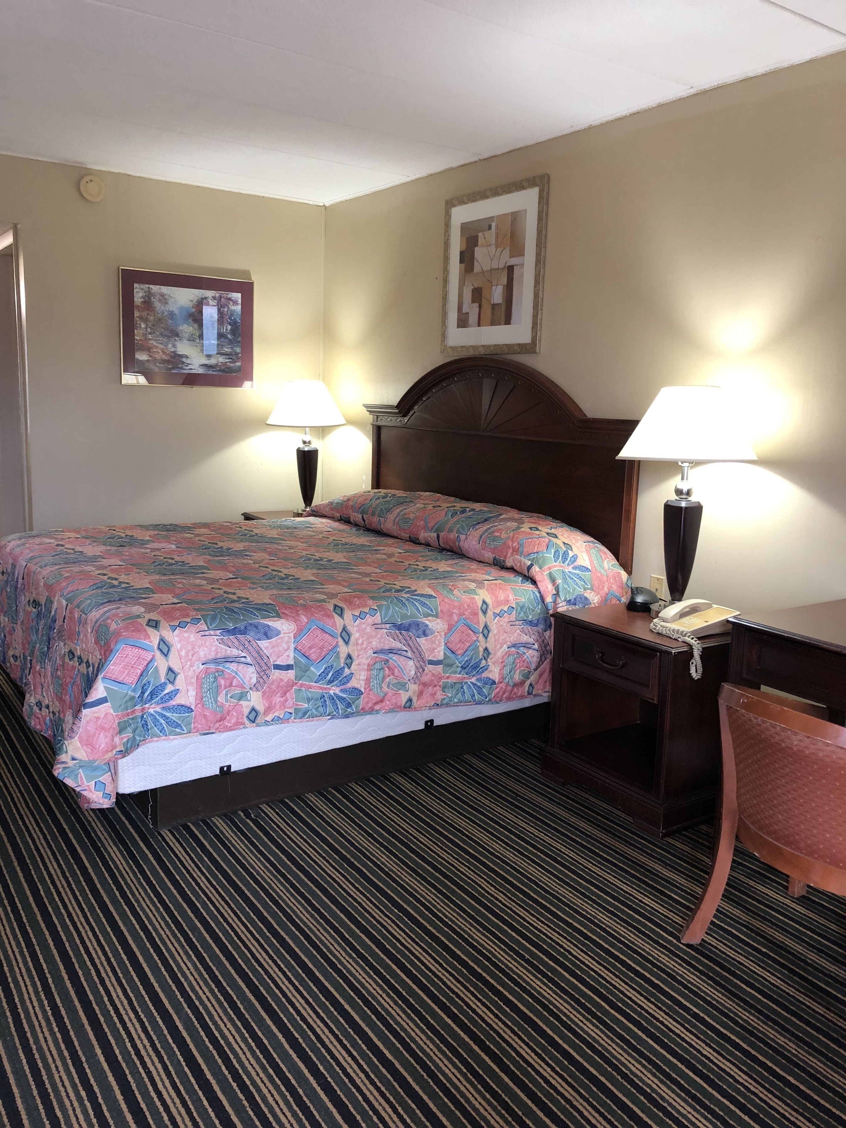 A hotel room with a bed, two bedside lamps, a desk with a telephone, a chair, and two framed pictures on the wall.