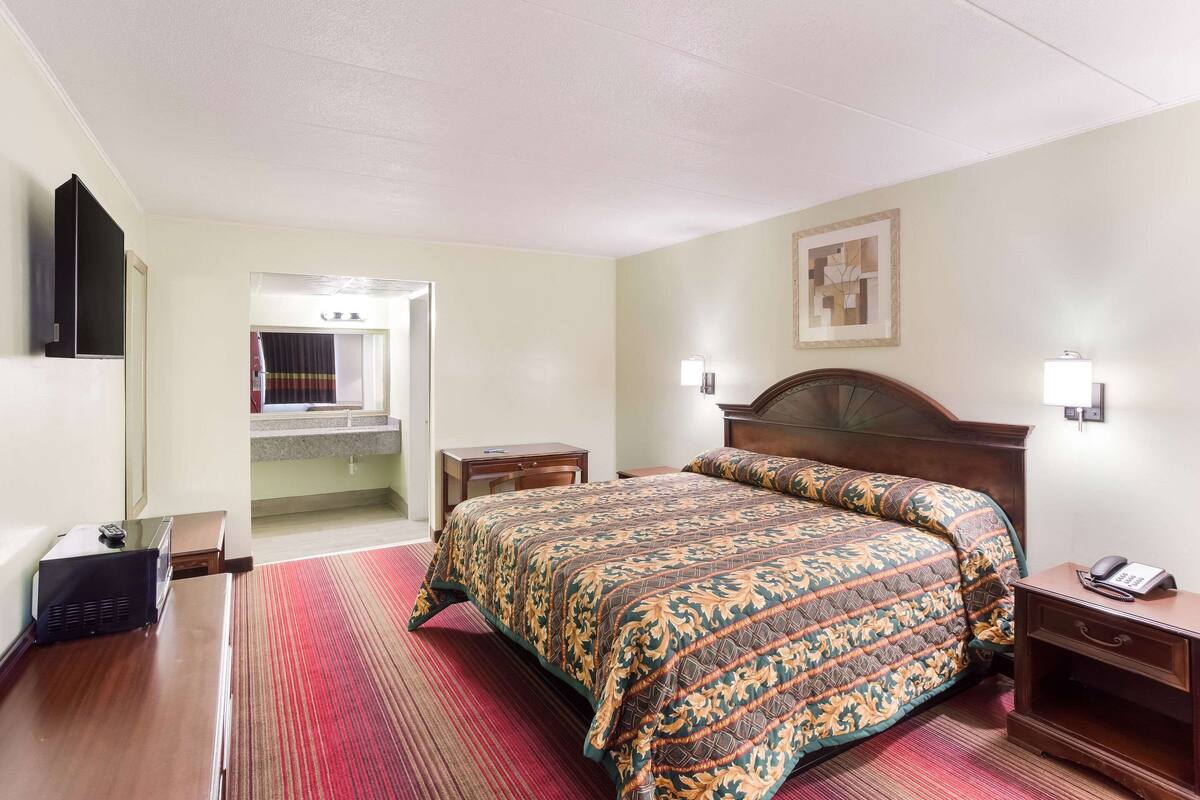 A hotel room with a large bed, a wooden nightstand, a bedside table, a mirror, and a door leading to a bathroom.