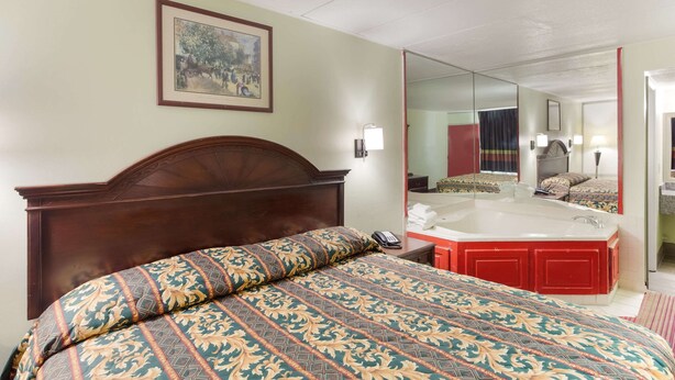 A hotel room with a large bed, a wooden headboard, a painting on the wall, a mirror, a red bathtub, and a telephone on the nightstand. - Rodeway Inn South Point (South Point)