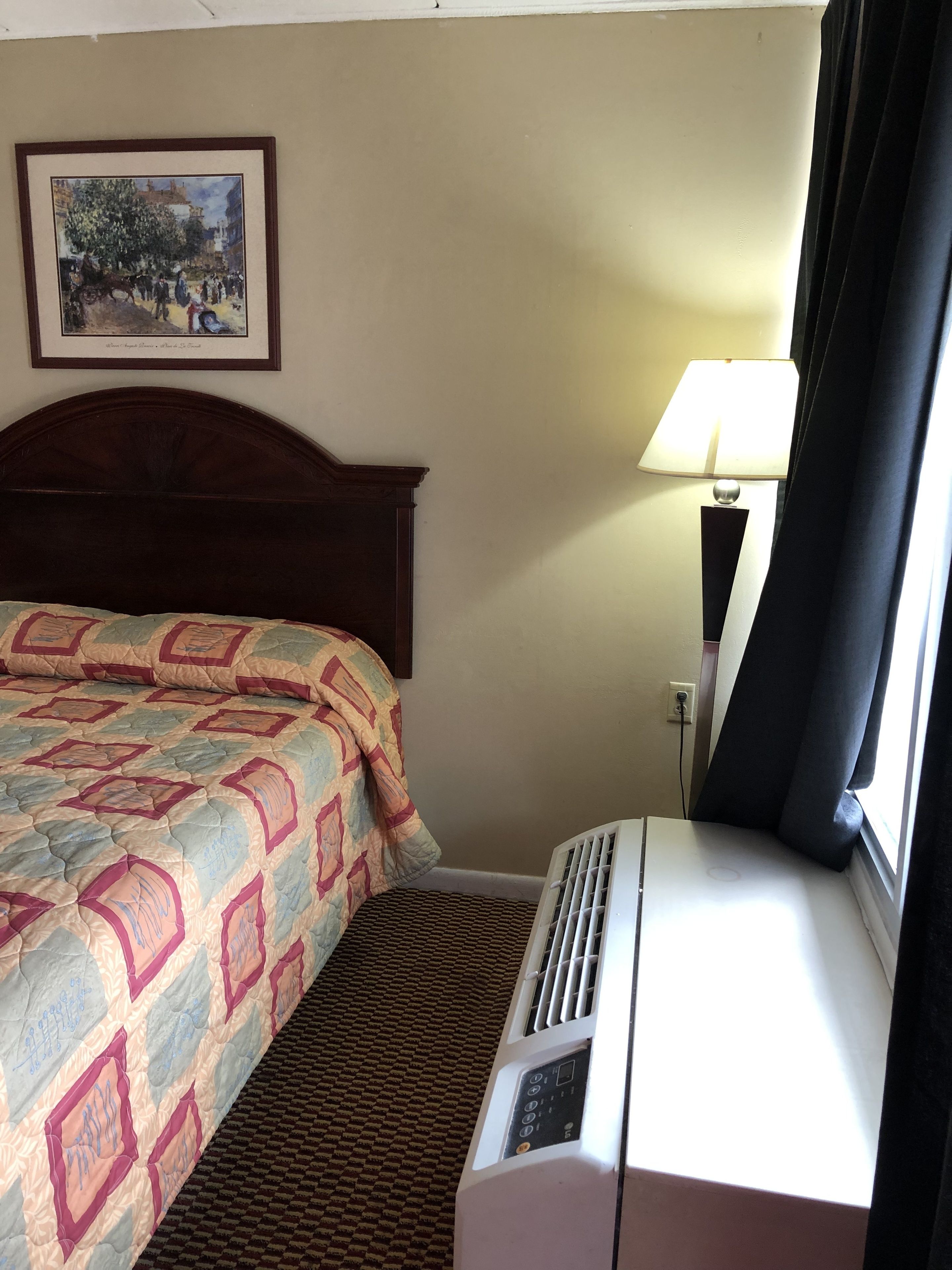 Habitación de hotel con cama, aire acondicionado de pared, lámpara y un cuadro enmarcado.