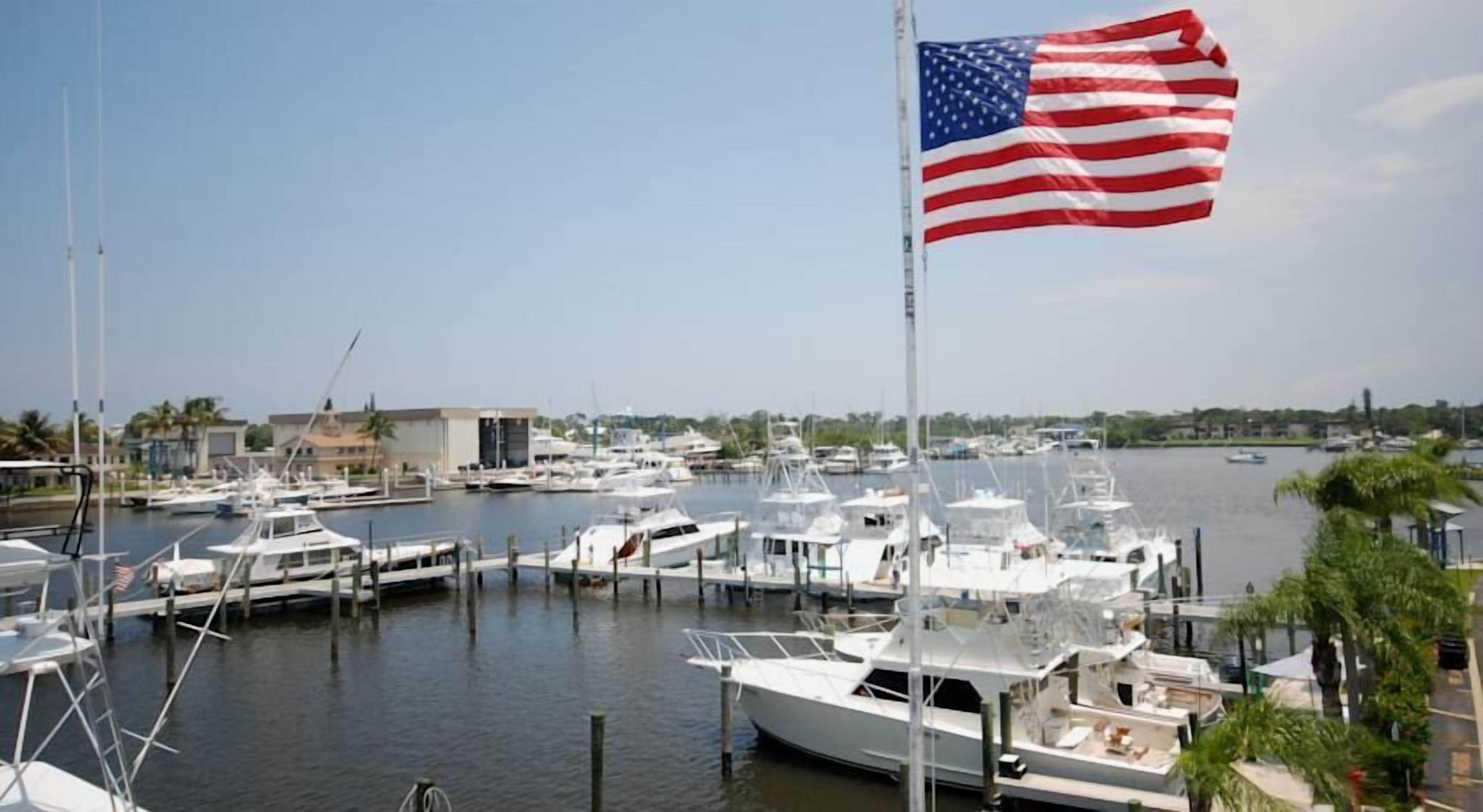 A marina with multiple boats docked, a flagpole with an American flag, and buildings in the background.
