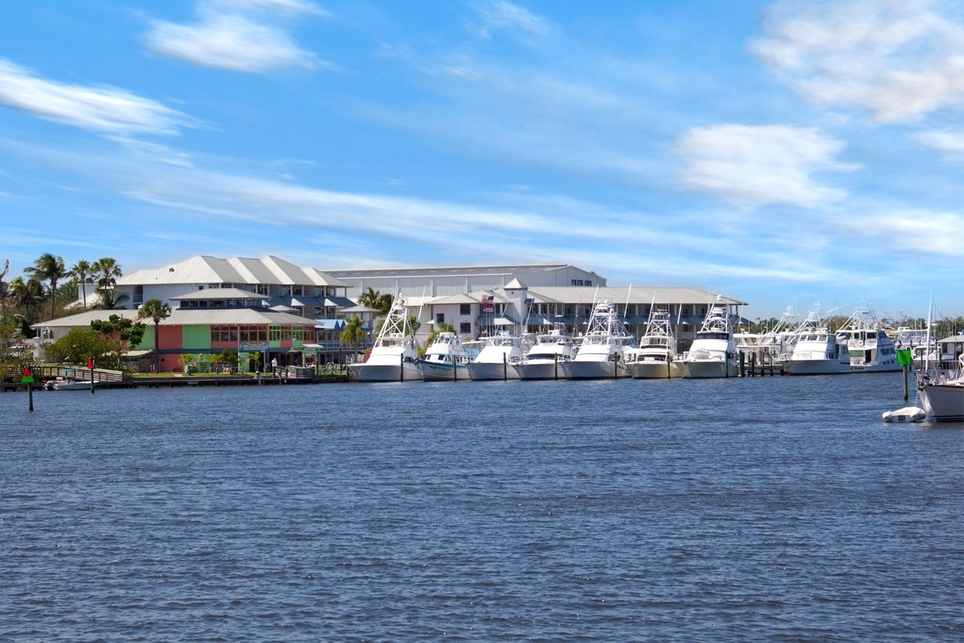 Un puerto deportivo con varias embarcaciones amarradas, un edificio grande con techo plano y un cielo azul despejado.